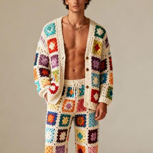 Op de afbeelding: Een crèmekleurig gehaakt vest en bijpassende broekset. Het vest heeft een knoopsluiting en de broek heeft een taille met trekkoord. Beide stukken hebben kleurrijke vierkante patronen in verschillende kleuren, waaronder rood, blauw, groen en geel.