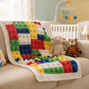 Puede incluir: Una manta de bebé hecha a mano con un diseño de bloques en rojo, azul, verde y amarillo. La manta está sobre un sofá crema, con peluches. El entorno de la guardería incluye una cuna y un móvil.