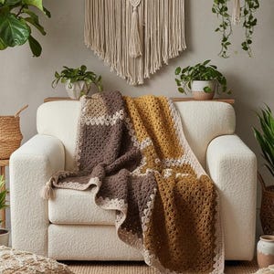 Op de afbeelding: Een gezellige woonkamer met een crèmekleurige bank, gedrapeerd met een handgemaakte gehaakte deken in bruin- en goudtinten. Een macramé wandkleed en potplanten versterken de bohemien stijl. Decoratieve kussens en een geweven vloerkleed maken de uitnodigende sfeer compleet.