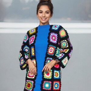 May include: A woman wearing a blue dress and a colorful crochet cardigan. The cardigan is made of black yarn with colorful squares.