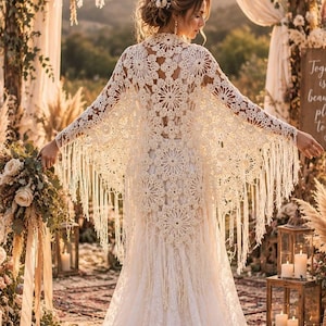 Puede incluir: Un vestido de novia de encaje color marfil hasta el suelo con una capa de ganchillo con grandes motivos circulares y largos flecos a lo largo de las mangas y el dobladillo. La prenda tiene un corpiño ajustado y una falda fluida.