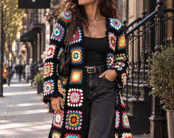 May include: A long-sleeved cardigan made of black yarn featuring a colorful granny square pattern in shades of red, yellow, blue, pink, and white. The garment is worn over a black tank top and black denim jeans with a black belt and black shoes.