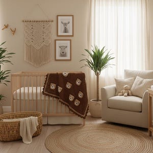 May include: A dark brown crochet blanket featuring repeating bear face motifs, draped over a light wood crib. The blanket has a scalloped edge and is positioned in a room with a macrame wall hanging, a beige armchair, and a woven floor basket.