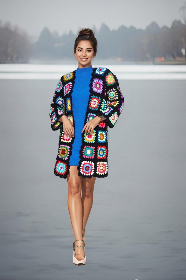 May include: A woman wearing a blue dress and a colorful crochet cardigan. The cardigan is made of black yarn with colorful squares.