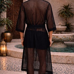May include: A black mesh robe with a matching belt. The robe has a knee-length design with three-quarter sleeves. The person is standing in front of a fountain and a stone wall.