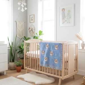 May include: A light blue crochet baby blanket featuring a grid pattern of six tan bear faces with dark brown noses and eyes, finished with a tan border, draped over the side of a light-colored wooden crib with vertical slats.
