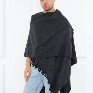 May include: A dark gray fringed shawl with a textured weave. The shawl is draped around a person's shoulders and is worn with light blue jeans.