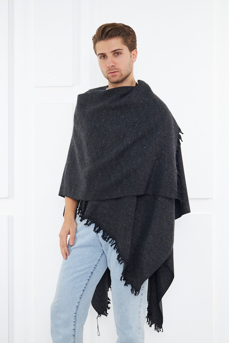 May include: A dark gray fringed shawl with a textured weave. The shawl is draped around a person's shoulders and is worn with light blue jeans.