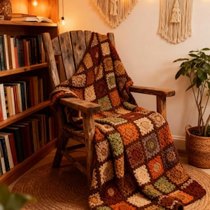 Puede incluir: Una manta de ganchillo hecha a mano, multicolor, sobre una silla de madera. La manta presenta un diseño de patchwork con cuadrados marrones, naranjas, verdes y crema. La silla está sobre una alfombra redonda de yute, con una estantería y tapices de macramé en el fondo.