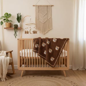 May include: A dark brown crochet baby blanket featuring a repeating pattern of square patches, each adorned with a small, light brown bear face appliqu&eacute;. The blanket has a decorative scalloped border and is draped over a light wood slatted crib.