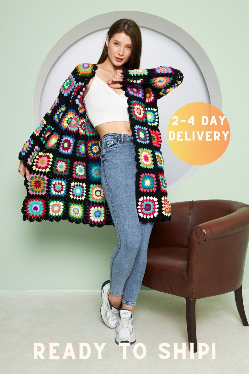 May include: A woman wearing a long, colorful crochet cardigan with a black background and a white tank top. She is also wearing blue jeans and white sneakers. The cardigan has a variety of colors, including red, green, blue, yellow, and pink. The background is a light green color.