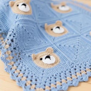 May include: A light blue crochet blanket featuring a repeating pattern of square motifs. Each square contains a beige bear face with a white muzzle and black nose. The blanket has a decorative scalloped border in light blue and beige yarn.
