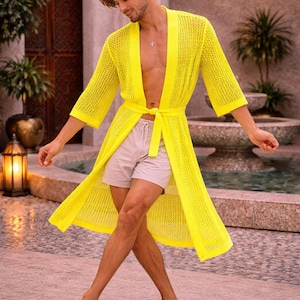 May include: A bright yellow, open-weave knit robe with a matching tie belt. The robe has three-quarter sleeves and is worn with light-colored shorts. The image is taken outdoors, with a fountain and greenery in the background.