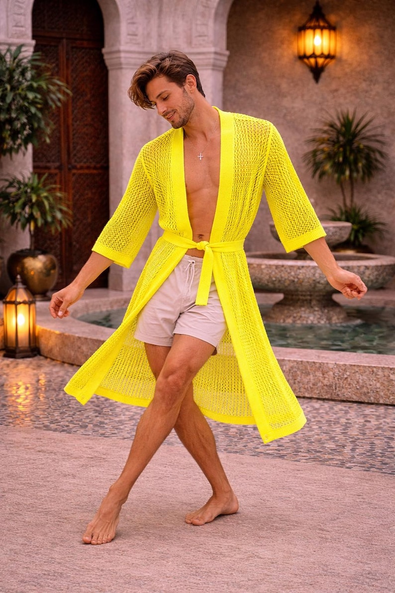 May include: A bright yellow, open-weave knit robe with a matching tie belt. The robe has three-quarter sleeves and is worn with light-colored shorts. The image is taken outdoors, with a fountain and greenery in the background.