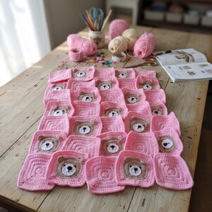 May include: A handmade crochet blanket composed of multiple square motifs in light pink yarn. Several squares feature a centered, circular bear face applique in tan and white yarn with black embroidered details for the eyes and nose.