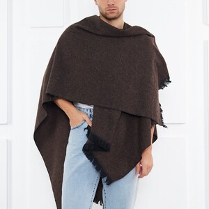 May include: A brown fringed shawl with a textured pattern. The shawl is draped around a person's shoulders and is worn with light blue jeans.