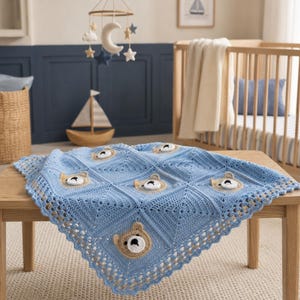 May include: A light blue crochet baby blanket featuring a repeating square pattern with centered, three-dimensional tan and white bear faces. The blanket has a decorative scalloped border and is constructed from yarn in a textured stitch.