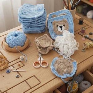 May include: A collection of light blue crochet squares, including one with a tan bear face design, alongside a ball of light blue yarn, white yarn, scissors, buttons, and measuring tapes arranged on a wooden craft table with open drawers.