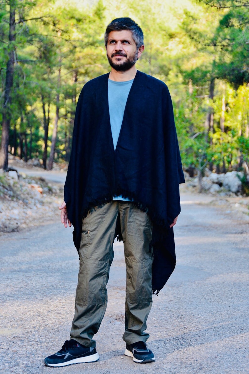 May include: A man wearing a dark blue poncho with a fringed trim, standing on a paved path in a woodland setting. He is wearing khaki cargo trousers and black trainers.