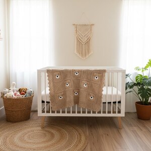 May include: A light brown crocheted baby blanket featuring a repeating pattern of bear faces with dark eyes and noses, draped over a white wooden crib. The blanket is constructed from square motifs with a textured border and neutral tones.