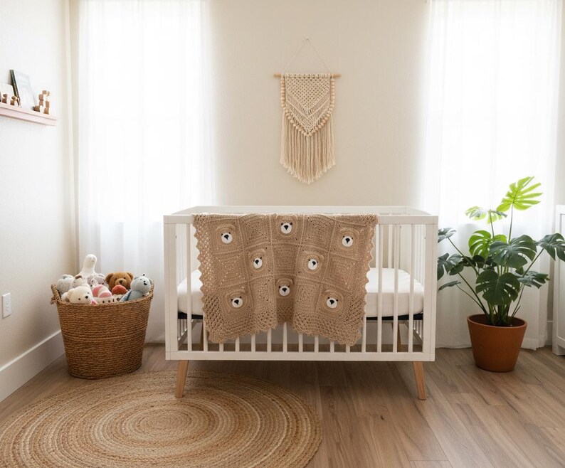 May include: A light brown crocheted baby blanket featuring a repeating pattern of bear faces with dark eyes and noses, draped over a white wooden crib. The blanket is constructed from square motifs with a textured border and neutral tones.