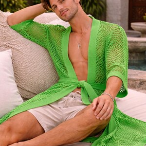 May include: A bright green, open-weave knit robe with a tie waist. The robe is worn over beige shorts. The model is sitting on a beige cushion. A silver cross necklace and bracelet are visible.