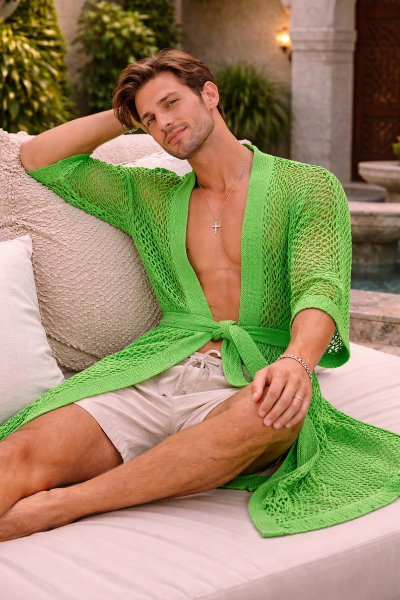 May include: A bright green, open-weave knit robe with a tie waist. The robe is worn over beige shorts. The model is sitting on a beige cushion. A silver cross necklace and bracelet are visible.