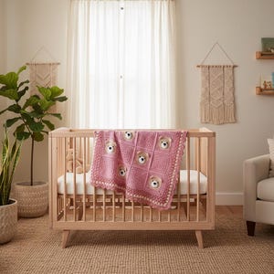 May include: A light pink crocheted baby blanket featuring a repeating square pattern with integrated plush bear faces. The blanket has a decorative scalloped border and is draped over the side of a light-toned wooden crib with vertical slats.