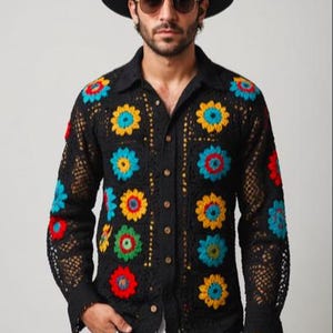 May include: A black crochet shirt with colourful floral designs. The shirt has a button-up front and long sleeves. The model is wearing a black hat with a blue and red band.