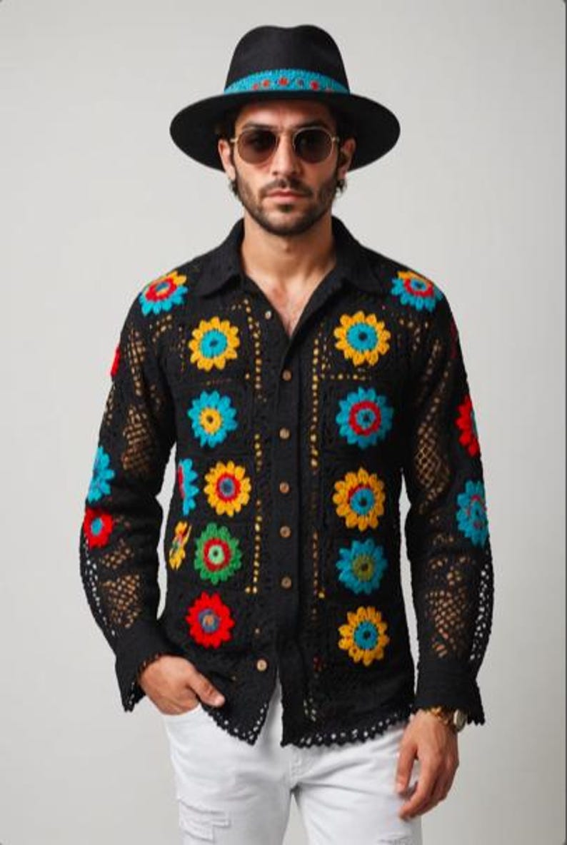 May include: A black crochet shirt with colourful floral designs. The shirt has a button-up front and long sleeves. The model is wearing a black hat with a blue and red band.
