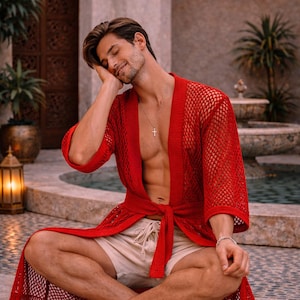 May include: A red mesh robe with a tie waist. The robe is worn over beige shorts. A silver cross necklace and bracelet are visible. The person is sitting cross-legged.