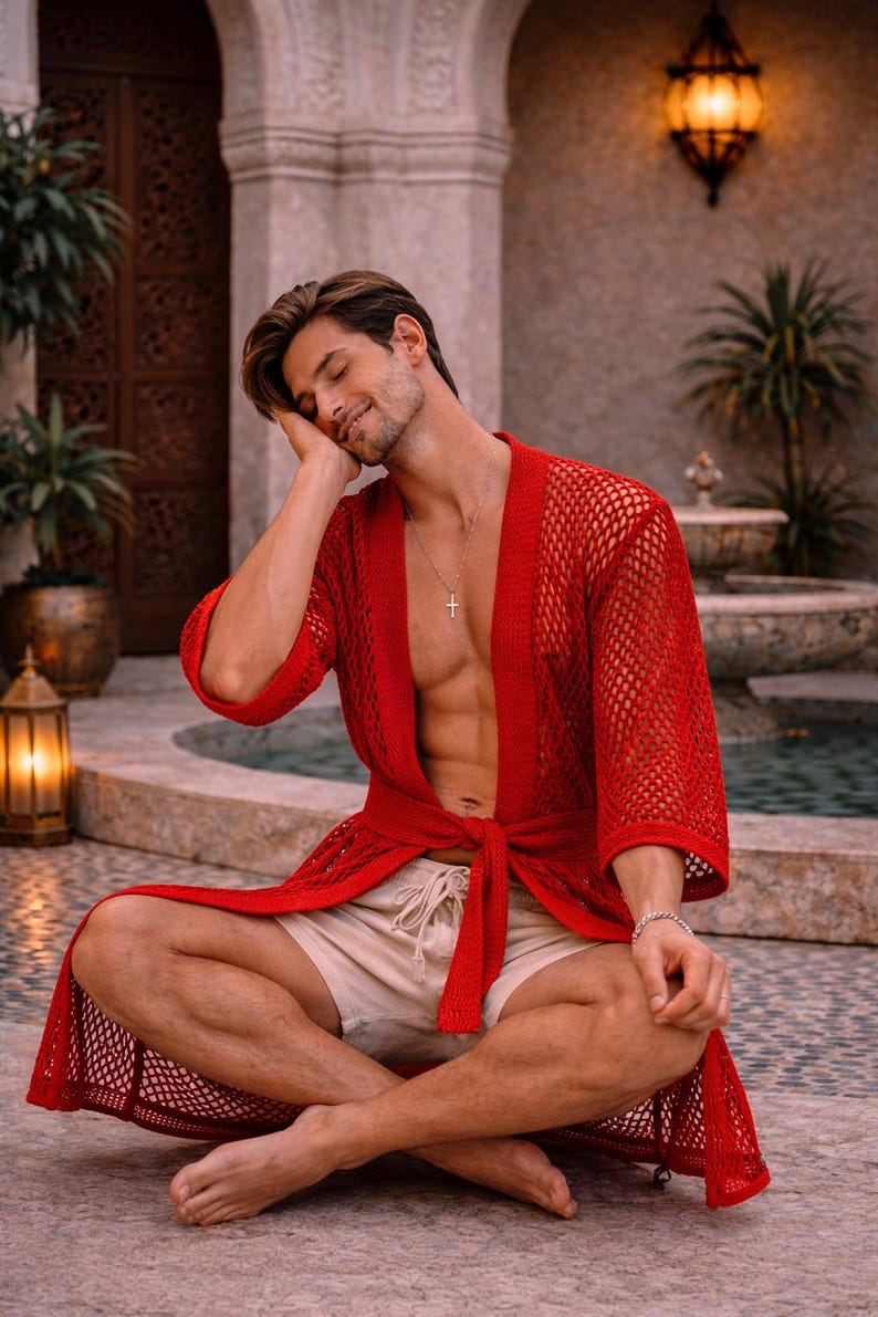 May include: A red mesh robe with a tie waist. The robe is worn over beige shorts. A silver cross necklace and bracelet are visible. The person is sitting cross-legged.