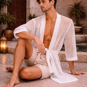May include: A white, open-weave mesh robe with a tie waist. The robe is paired with beige shorts. The image is set outdoors with a stone floor and architectural details in the background.