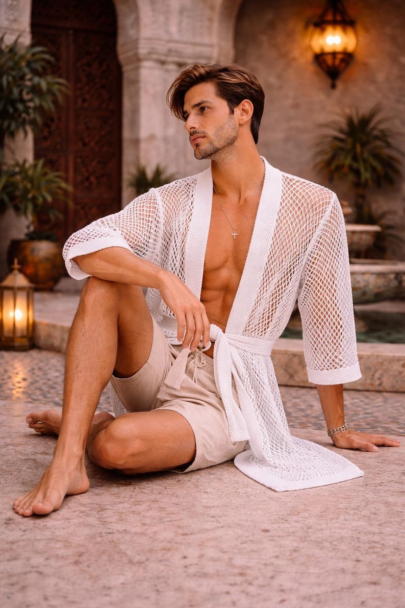 May include: A white, open-weave mesh robe with a tie waist. The robe is paired with beige shorts. The image is set outdoors with a stone floor and architectural details in the background.