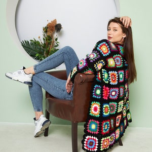 May include: A woman sits in a brown chair wearing a black crochet cardigan with colorful squares. She is wearing blue jeans and white sneakers.