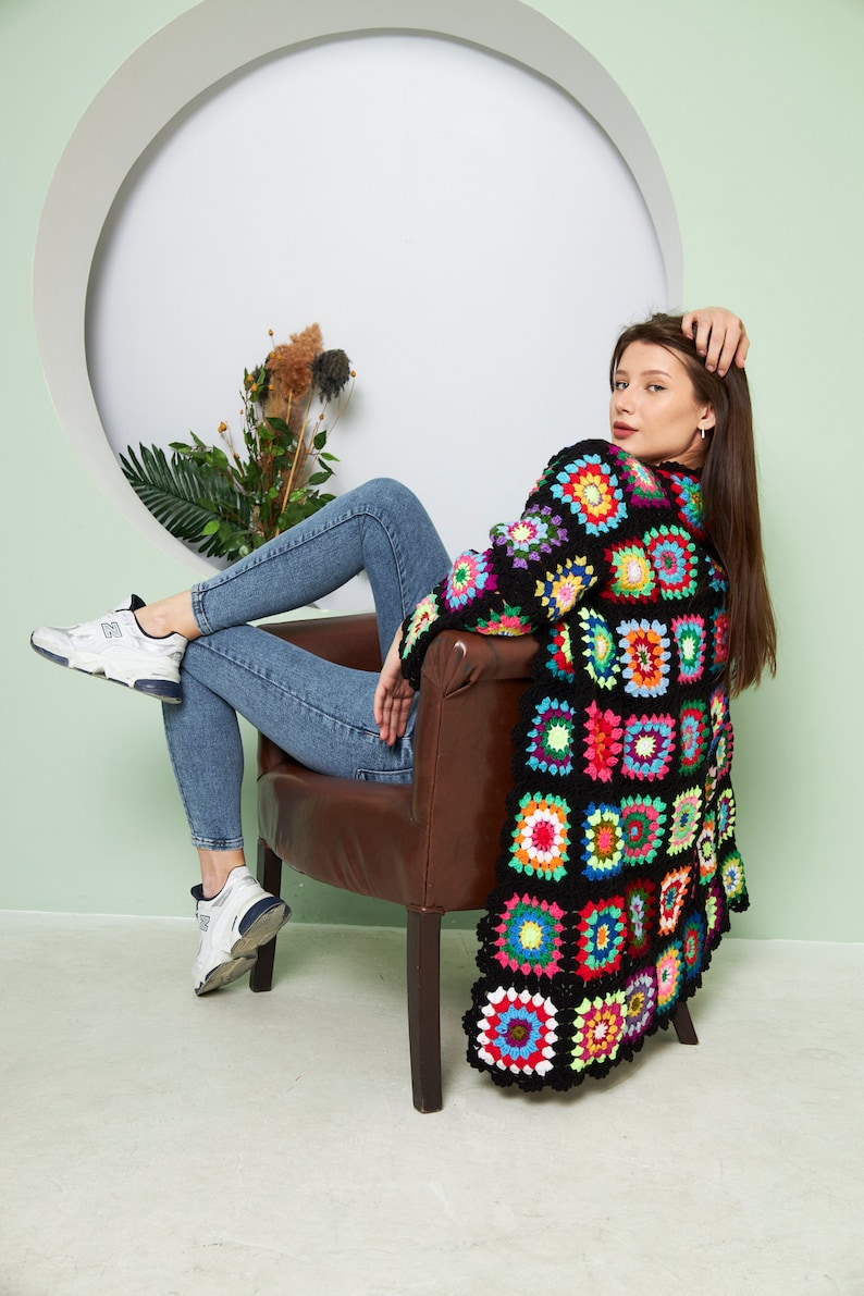 May include: A woman sits in a brown chair wearing a black crochet cardigan with colorful squares. She is wearing blue jeans and white sneakers.