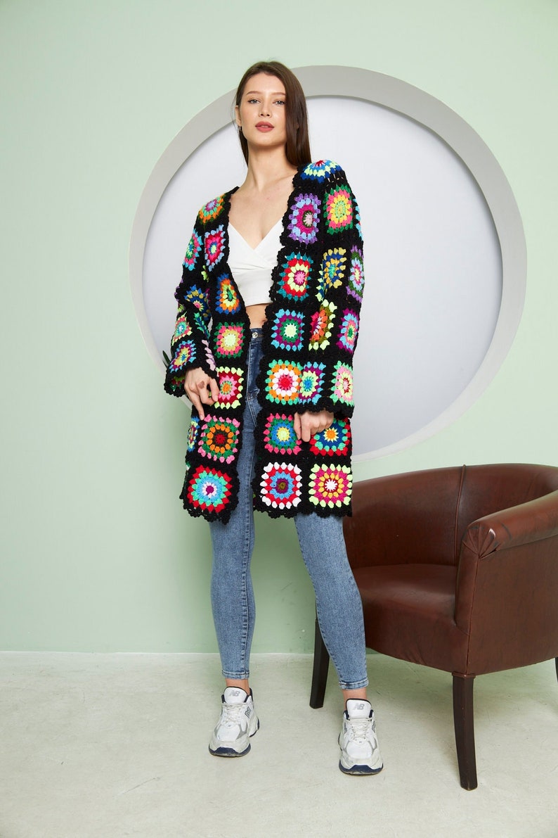 May include: A long black crochet cardigan with colorful granny squares. The cardigan is open in the front and has long sleeves. The model is wearing blue jeans and white sneakers.