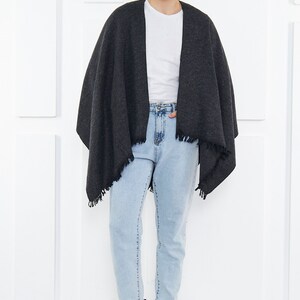 May include: A person wearing a dark gray shawl with a fringe trim over a white t-shirt and light blue jeans. They are wearing black leather boots.