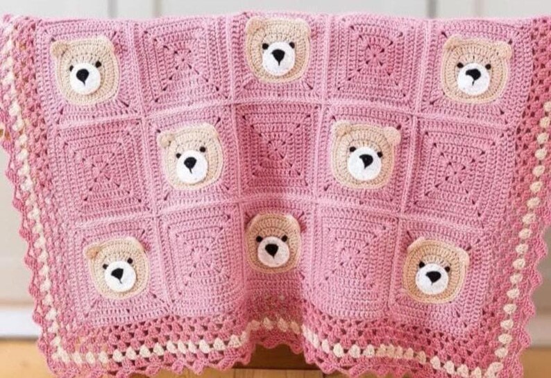May include: A pink crochet blanket featuring a repeating pattern of square blocks. Several blocks contain a beige bear face with a white muzzle and black nose. The blanket is finished with a decorative scalloped border in pink and cream yarn.