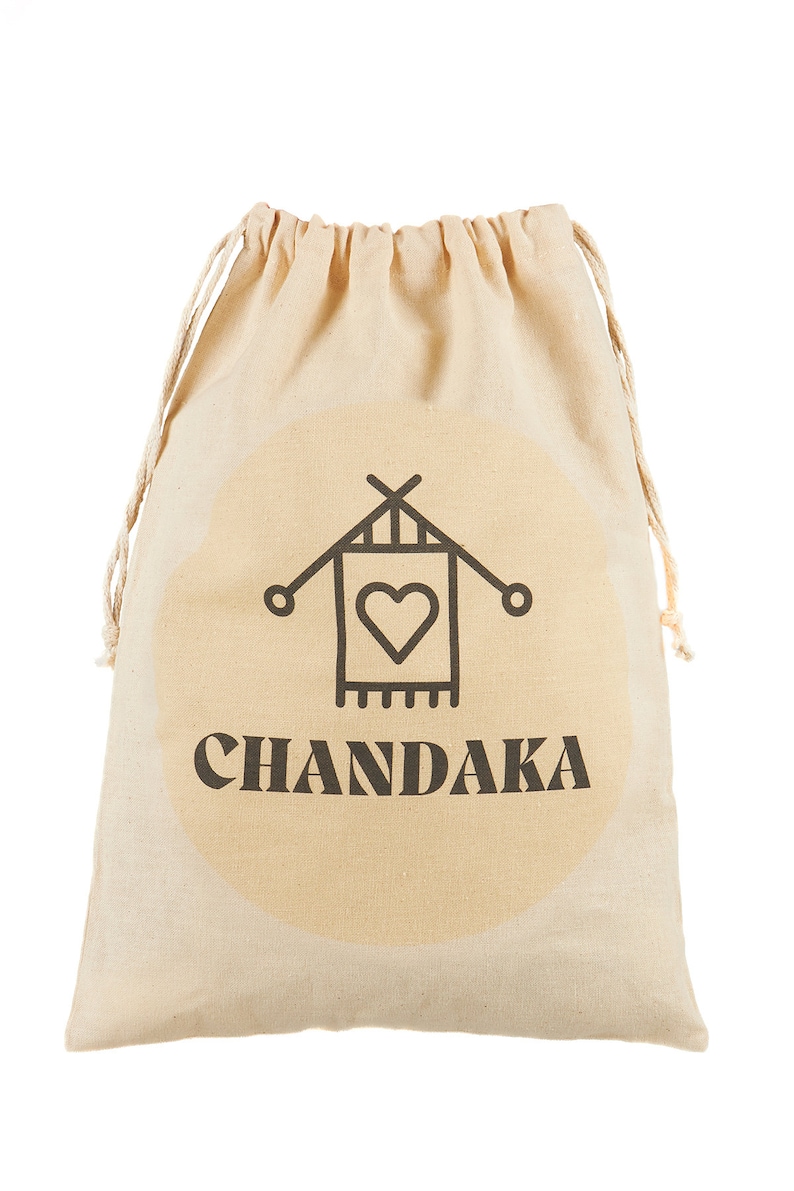 May include: Beige drawstring bag with a circular design and the word "CHANDAKA" printed in dark brown. The bag features a black graphic of a house with a heart inside. The drawstring is a light beige color.