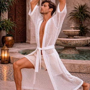 May include: A white, open-weave robe with a matching belt is worn over beige shorts. The robe has a long, flowing design. The person is in a yoga pose with arms raised. The background includes a fountain and lush greenery.