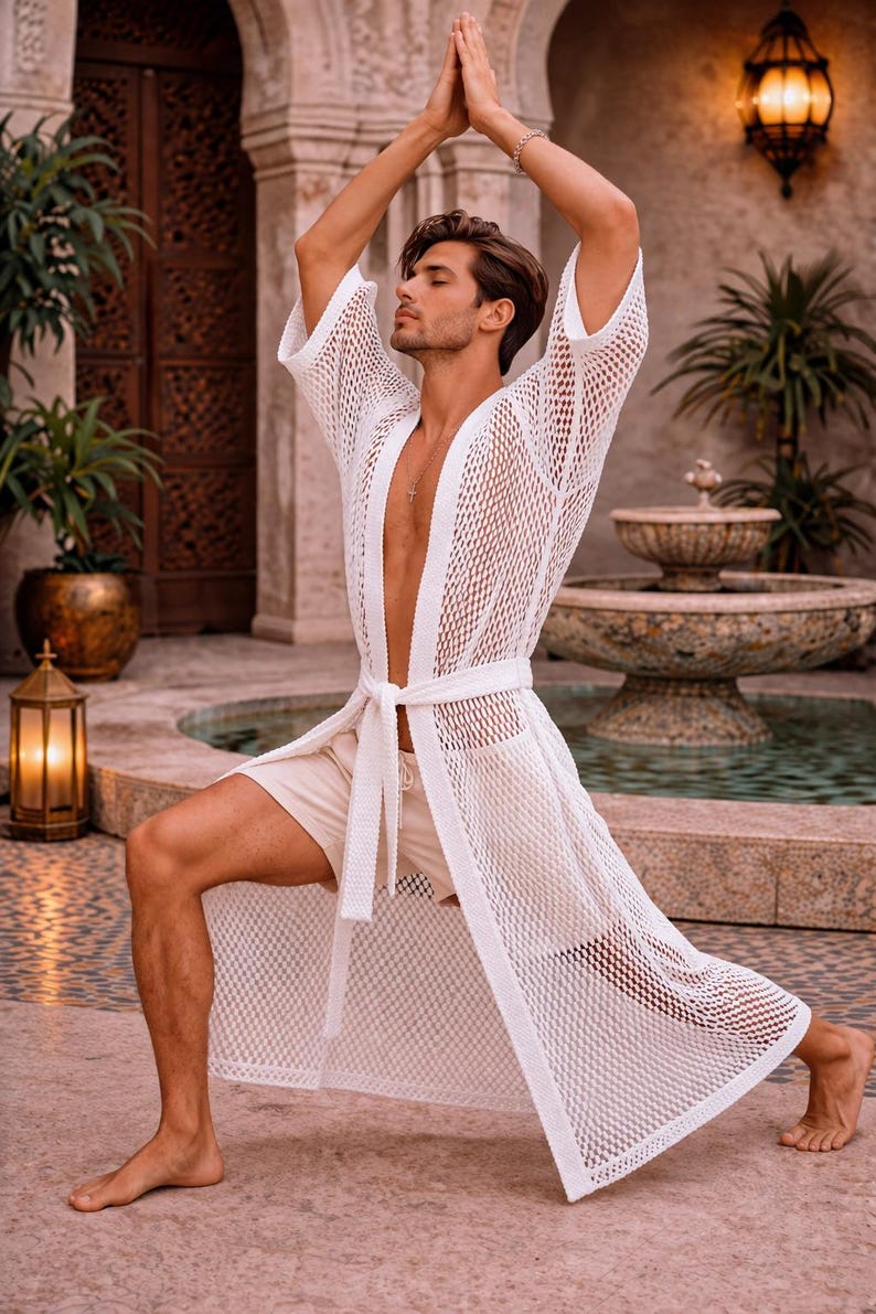 May include: A white, open-weave robe with a matching belt is worn over beige shorts. The robe has a long, flowing design. The person is in a yoga pose with arms raised. The background includes a fountain and lush greenery.