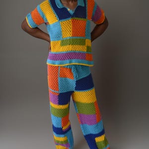 May include: A colourful crochet outfit with a short-sleeved shirt and trousers. The shirt and trousers are made of a patchwork of different coloured squares. The outfit is a bright mix of orange, yellow, green, blue, and purple.