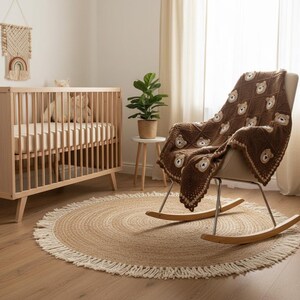 May include: A dark brown crochet blanket featuring a repeating pattern of light brown bear faces in square sections, finished with a decorative scalloped border. The blanket is draped over a rocking chair with a light-colored seat and wooden rockers.