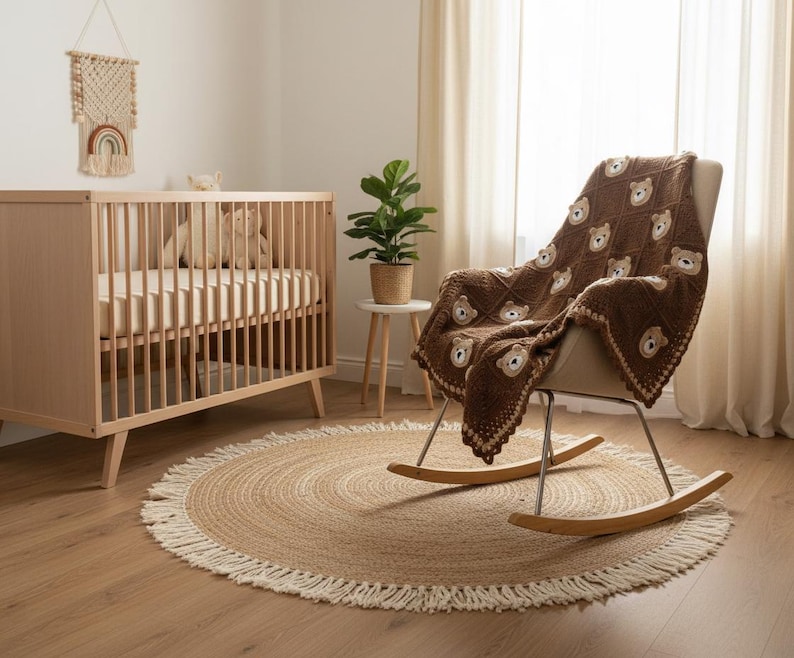 May include: A dark brown crochet blanket featuring a repeating pattern of light brown bear faces in square sections, finished with a decorative scalloped border. The blanket is draped over a rocking chair with a light-colored seat and wooden rockers.