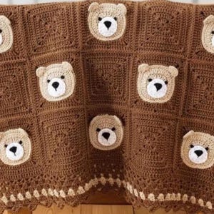 May include: A crochet blanket made of dark brown square panels, each featuring a centered beige bear face with a black nose and eyes. The blanket has a decorative border with a repeating pattern of small beige squares along the bottom edge.