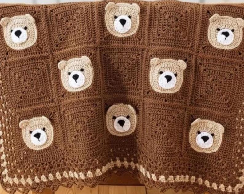 May include: A crochet blanket made of dark brown square panels, each featuring a centered beige bear face with a black nose and eyes. The blanket has a decorative border with a repeating pattern of small beige squares along the bottom edge.