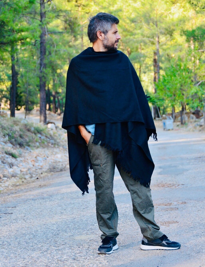 May include: A person wearing a black poncho and green cargo trousers. The person is standing on a paved road with trees in the background.