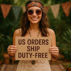 May include: A smiling person wearing sunglasses and a headband holds a sign with the text "US ORDERS SHIP DUTY-FREE". The person is wearing a cream-colored dress and jewelry. The background includes greenery and colorful pennant flags.