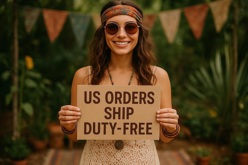 May include: A smiling person wearing sunglasses and a headband holds a sign with the text "US ORDERS SHIP DUTY-FREE". The person is wearing a cream-colored dress and jewelry. The background includes greenery and colorful pennant flags.